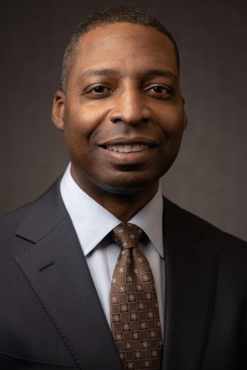 Mr. James Heffner, an African American Male wearing a grey suit jacket with a white collared shirt and a red tie with gold dots.